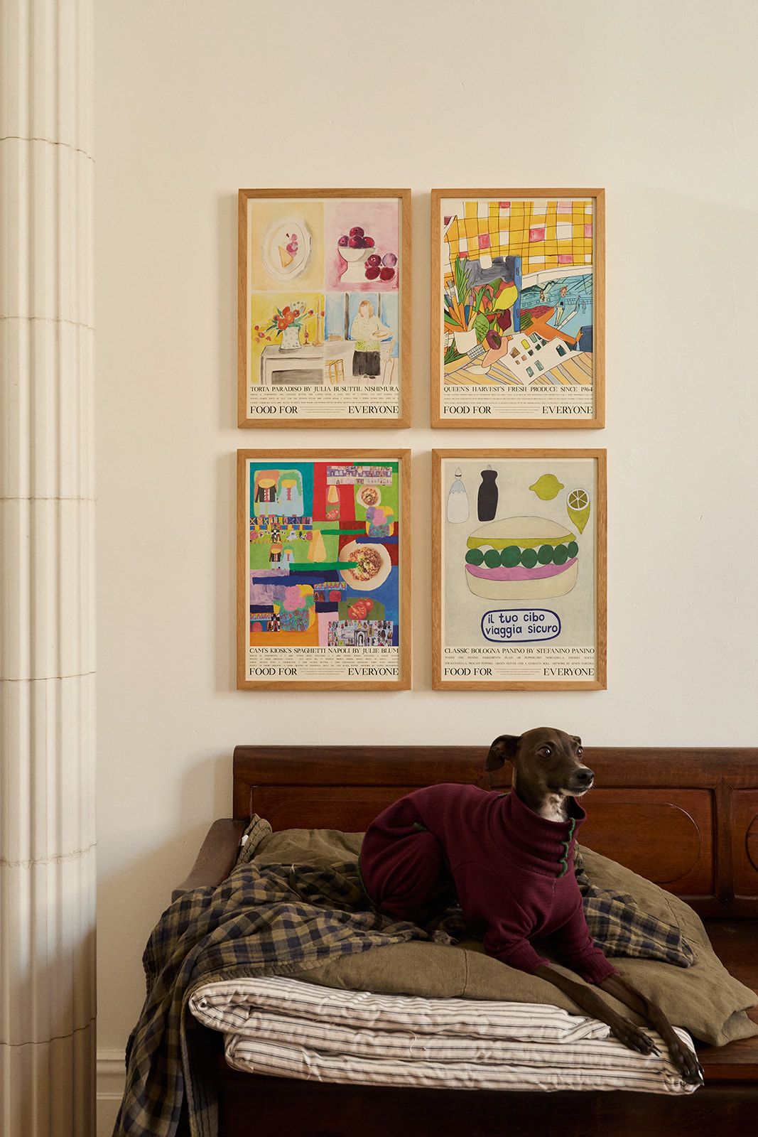 A photo of four Food For Everyone posters on display in frames hanging upon a wall. In front of the framed posters is a brown couch with a greyhound dog sitting on it.